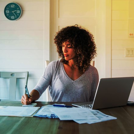 A woman sits at her dining room table with laptop and financial reports doing her monthly budget. She is writing down budgets as she works on her computer to do monthly finances, pay taxes and save money for the future.