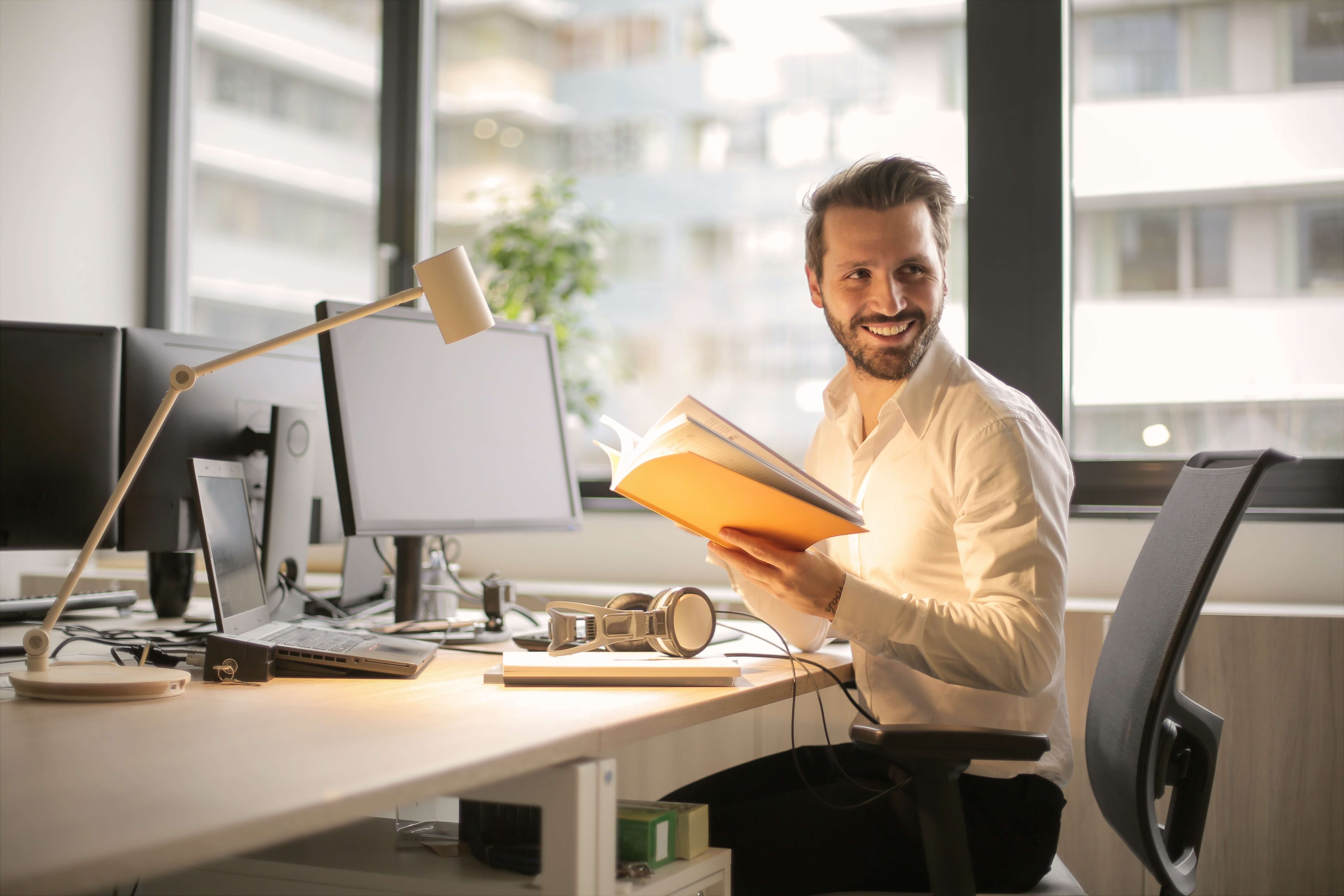 Smiling man holding yellow book, turns around in desk seat and looks back